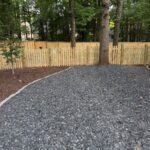 A light wood picket fence borders a yard filled with grey gravel and dark mulch.